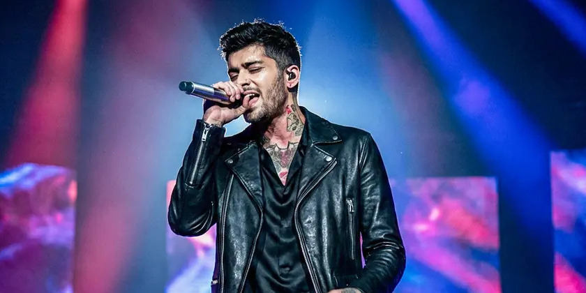 ZAYN holds a microphone to his mouth in purple lighting.