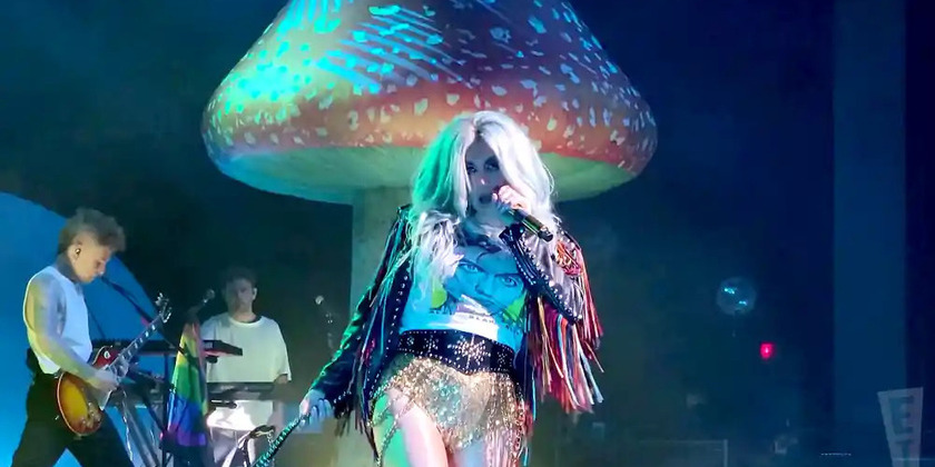Kesha wearing a fringe jacket, sparkly shorts, and a graphic tee while performing on stage.