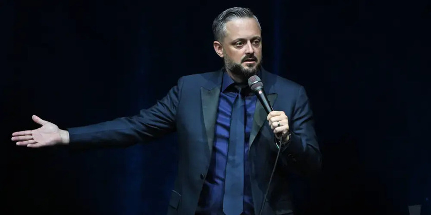 Nate Bargatze holds his hand out to the side while wearing a suit and performing stand-up.