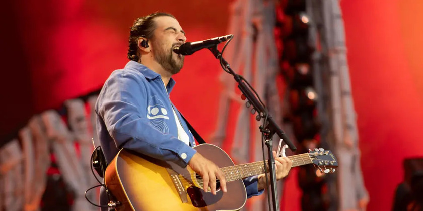 Noah Kahan holds a guitar on stage and sings.