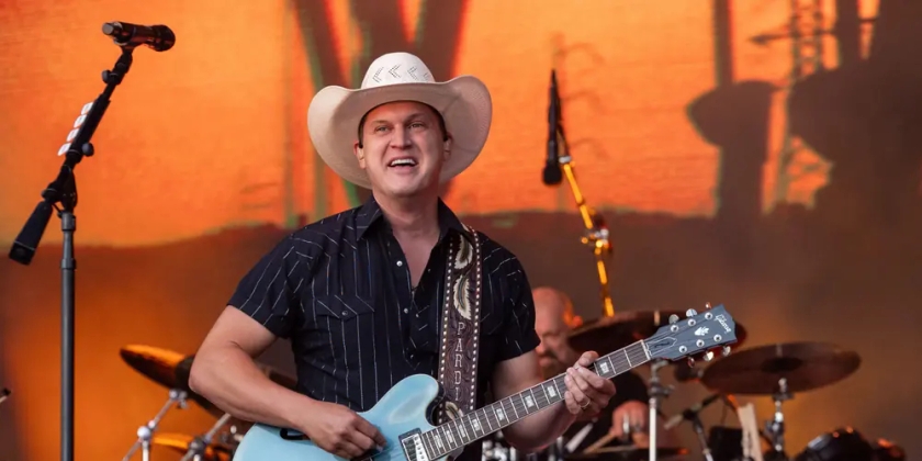 Jon Pardi smiles while singing on stage, a blue guitar in his hand.