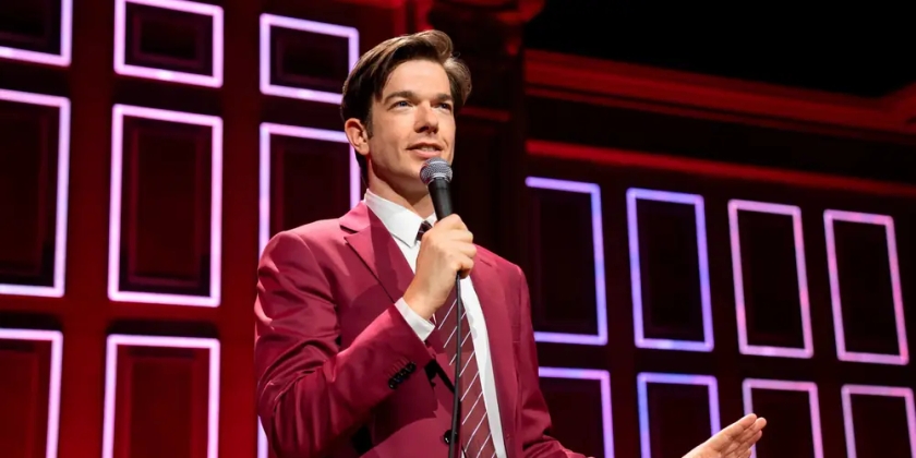 John Mulaney wears a red suit and tells a joke on stage during a stand-up set.