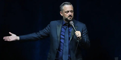 Nate Bargatze holds his hand out to the side while wearing a suit and performing stand-up.