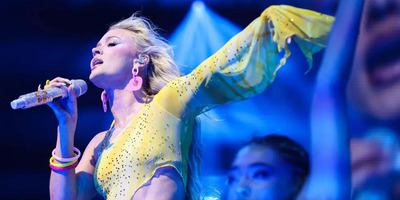 Zara Larsson sings in a yellow sparkly outfit with her hand in the air and hair blowing.