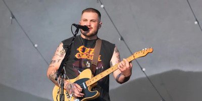 Zach Bryan sings on stage with a guitar in a band t-shirt.