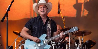 Jon Pardi smiles while singing on stage, a blue guitar in his hand.
