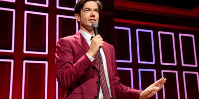 John Mulaney wears a red suit and tells a joke on stage during a stand-up set.