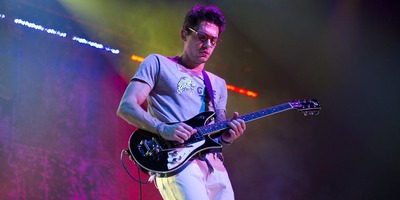 John Mayer plays guitar on stage under colorful lighting.